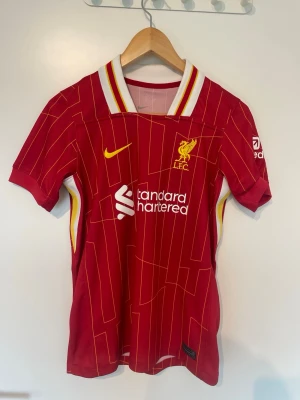 Liverpool matchtröja röd Nike - Säljer en röd Liverpool matchtröja från Nike med gula och vita detaljer. Tröjan har klubbmärke och sponsortryck på bröstet, samt vita och gula ränder på kragen. Perfekt för dig som älskar fotboll och vill stötta ditt lag på match eller träning. 
