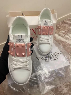 Vita sneakers med rosa nitar - Snygga vita sneakers från Valentino med unika rosa detaljer och pyramidformade nitar över snörningen. Skorna har en låg profil och är tillverkade i slätt läder med vita skosnören. Perfekta för dig som vill sticka ut med en cool och trendig look.
