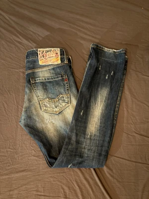 Blå slitna jeans från Replay  - Snygga blå jeans från replay med slitningar och ljusa partier på benen. Klassisk femficksmodell med raka ben och detaljerade sömmar. Jeansen har en cool vintage-look och patch på bakfickan. Perfekta för en avslappnad streetstil.