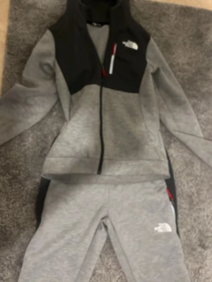 Grått loungewear-set från The North Face - Snyggt och bekvämt loungewear-set från The North Face i ljusgrått med svarta detaljer. Setet består av en zip-hoodie med fickor och matchande byxor med logga på låret. Perfekt för chill dagar hemma eller när du vill vara casual. Mjukt material och modern passform.