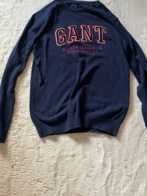 Mörkblå GANT tröja  - Mörkblå långärmad tröja från GANT med röd och vit text på bröstet där det står 'GANT New Haven Connecticut'. Klassisk rund halsringning och mjukt material, perfekt för en avslappnad stil.