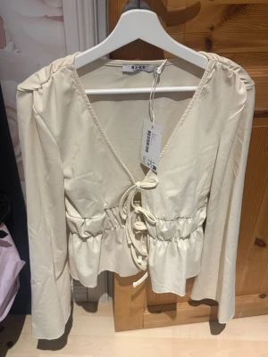 Beige omlottblus med snörning från NA-KD - Trendig beige omlottblus från NA-KD med långa ärmar och snygg snörning framtill. Blusen har v-ringning och markerad midja med volangdetalj. Perfekt att styla med jeans eller kjol för en chic look.