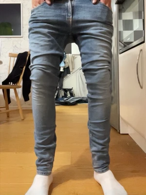 Nudie slim fit  - Säljer ett par ljusblå slim fit jeans med klassisk femficksdesign och dragkedjegylf. Jeansen har diskreta slitningar och kontrastsömmar på bakfickorna. Perfekta för en avslappnad och trendig look.