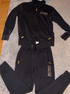 Svart loungewear-set från BOSS - Snyggt svart loungewear-set från BOSS med gulddetaljer på dragkedjor och logga på både tröja och byxa. Setet består av en zip-hoodie och matchande byxor med resår och dragsko i midjan. Perfekt för chill dagar hemma. Storlek S/M