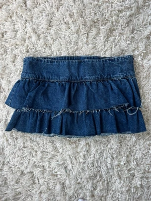Blå volangkjol i denim från Gina Tricot - Säljer en blå kort jeanskjol från Gina Tricot med dubbla volanger och råa kanter för en trendig look. Kjolen har en klassisk midja och är perfekt för dig som gillar denim och vill ha en cool stil.är aldrig använd!❤️