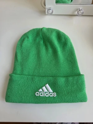 Grön mössa från Adidas - Snygg grön mössa från Adidas med uppvikt kant och broderad vit logga framtill. Tillverkad i mjukt, stretchigt material som sitter skönt på huvudet. Perfekt accessoar för att addera färg till din outfit.