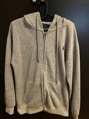Grå hoodie från Polo Ralph Lauren - Snygg grå zip-hoodie från Polo Ralph Lauren med klassisk huva och dragsko. Broderad logga på bröstet och två stora fickor framtill. Mjuk bomullsblandning som är perfekt för en chill och avslappnad stil.