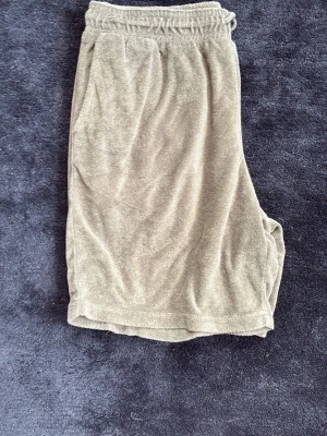 Beiga frottéshorts med snörning från John Henrik  - Mysiga beiga shorts i mjuk frotté med elastisk midja och snörning framtill. Shortsen har en relaxed passform och två sidofickor, perfekta för chill dagar eller sommarhäng.