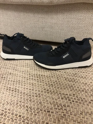 Mörkblåa BOSS skor - Snygga svarta sneakers från BOSS med vit sula och diskreta detaljer i grått. Ovandelen är i textil med stickad känsla och skorna har svarta skosnören. Perfekta för en stilren och modern look.