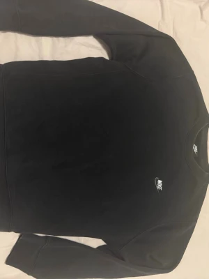 Svart sweatshirt från Nike - Klassisk svart sweatshirt från Nike med broderad vit logga på bröstet. Tröjan har rund halsringning och långa ärmar. Perfekt för en sportig och avslappnad stil.