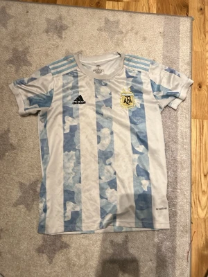 Argentina matchtröja Adidas blåvit - Säljer en Argentina matchtröja från Adidas med klassiska blå och vita ränder, AFA-emblem på bröstet och svarta Adidas-loggan. Tröjan har korta ärmar och är tillverkad i ett lätt, ventilerande material. Perfekt för fotbollsträning eller att bära till vardags.