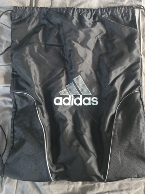 Svart ryggsäck från Adidas - Snygg svart ryggsäck från Adidas med klassisk logga i vitt framtill. Väskan har vita detaljer längs sidorna och är tillverkad i slitstarkt syntetmaterial. Perfekt för träning eller skola och har en sportig look.