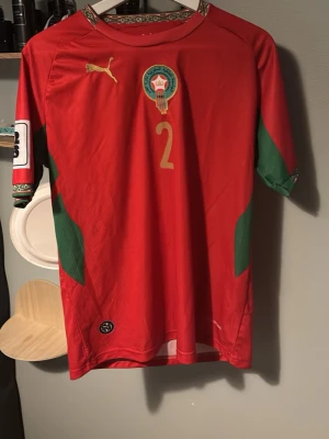 Marocko matchtröja Hakimi #2 Puma - Säljer en röd Marocko matchtröja från Puma med Hakimi och nummer 2 i guld på ryggen. Tröjan har gröna detaljer på sidor och ärmar, samt Marockos landslagsemblem på bröstet. Korta ärmar och sportig passform, perfekt för fotbollsfans.