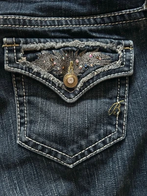 Mörkblå jeans med dekorerad ficka - Säljer ett par mörkblå jeans med snygga slitningar och kontrastsömmar. Bakfickan har ett unikt broderi med färgglada stenar och slitna detaljer, samt en dekorativ knapp. Jeansen har låg midja och klassisk femficksmodell. Perfekt för dig som gillar detaljer!