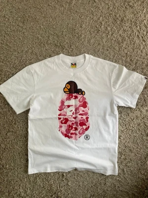 Vit BAPE t-shirt med rosa camo print - Säljer en vit t-shirt från BAPE med ikoniskt rosa camo-mönster och Baby Milo-apa på bröstet. Klassisk passform och rund hals. Perfekt för dig som gillar streetwear och vill sticka ut med en unik design.
