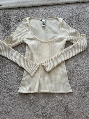 Ribbad beige långärmad topp - Snygg ribbad topp i beige med lång ärm och v-ringning. Toppen har en liten rosett framtill och är figurnära i modellen. Perfekt att styla med jeans eller kjol för en trendig look.