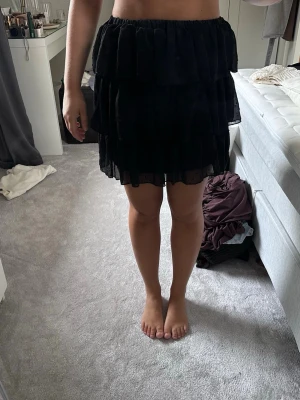Svart volangkjol i kort modell - Säljer en svart kort kjol med flera lager volanger. Kjolen har resår i midjan och är gjord i ett tunt, lätt material som ger ett flowy intryck. Perfekt för dig som gillar en feminin och trendig stil.