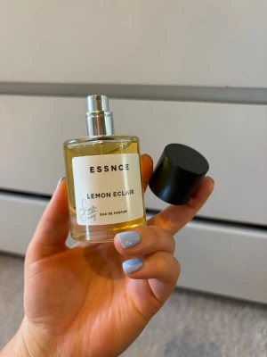 ESSNCE Lemon Eclair parfym - Fräsch parfym från ESSNCE med doften Lemon Eclair. Flaskan är cylindrisk i genomskinligt glas med svart rund kork och vit etikett. Innehållet är gulaktigt och flaskan har en enkel, modern design. Eau de Parfum-variant.
