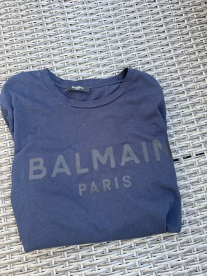 Mörkblå Balmain Paris t-shirt - Snygg mörkblå t-shirt från Balmain Paris med stor logga framtill. Klassisk rund halsringning och korta ärmar. Tillverkad i mjuk bomull som känns skön mot huden. Perfekt för dig som vill ha en stilren och trendig look.
