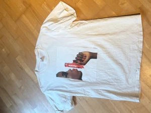 Vit t-shirt med Supreme-tryck - Säljer en vit t-shirt med ett stort Supreme-inspirerat tryck på framsidan som visar ett ansikte och en hand med en röd Supreme-bar. Klassisk passform och rund hals. Perfekt för dig som gillar streetwear och vill sticka ut.