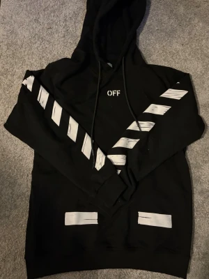 Off-WhiteHoodie - Säljer en svart hoodie med huva och dragsnören, prydd med vita diagonala ränder på ärmarna och stor grafisk print på ryggen' på bröstet. Hoodien har en streetwear vibe och är perfekt för dig som gillar statement-plagg.
