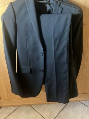 Klassisk svart kostym, tvådelad - Säljer en stilren svart kostym med kavaj och matchande byxor från dressman. Kavajen har två knappar, smal passform och klassiska fickor. Perfekt för dig som vill ha en enkel och tidlös look. Materialet känns mjukt och följsamt.