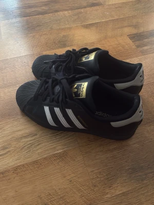 Adidas Superstar svarta sneakers - Klassiska Adidas Superstar sneakers i svart läder med vita ränder och gulddetaljer på plösen. Skorna har den ikoniska snäckformade tån och svart sula. Perfekta för dig som gillar retrostil och streetwear.