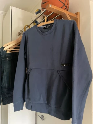 Mörkblå sweatshirt från Sail Racing - Mörkblå sweatshirt från Sail Racing med rund halsringning och ribbade muddar. Tröjan har en diskret logga på bröstet och en stor magficka framtill. Perfekt för en avslappnad och sportig stil.