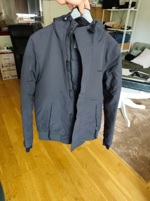 Mörkgrå vindjacka med huva - Snygg mörkgrå vindjacka med huva och dragkedja framtill. Jackan har två sidofickor med dragkedja och ribbade muddar vid ärmsluten. Tillverkad i ett lätt och vindtätt material, perfekt för blåsiga dagar.
