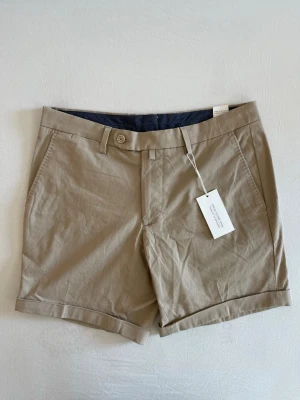 These Glory Days Chinosshorts - Säljer ett par sköna beiga chinosshorts från These Glory Days Slim Fit Artikelnummer: 72307510012 Size 48, W32 Fyra fickor med sleek design Knapp Stretch Herrshorts Nypris: 699kr