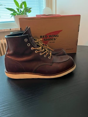 Bruna Red Wing boots i skinn - Säljer ett par klassiska bruna boots från Red Wing i skinn med vita sulor och gulbruna snören. Bootsens höga modell och robusta design gör dem till ett riktigt statement. Perfekta för dig som gillar tidlös stil och kvalitet.
