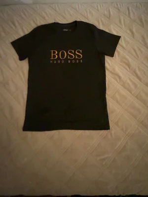 Svart Hugo Boss t-shirt med tryck - Snygg svart t-shirt från Hugo Boss med stort guldigt BOSS-tryck på bröstet. Klassisk passform och rund halsringning. Perfekt för dig som vill ha en stilren och trendig look.