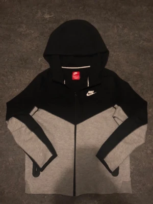 Nike Tech svart och grå - Nike tech i svart och grått med dragkedja framtill och klassisk Nike-logga på bröstet. Hoodien har huva och svarta detaljer längs ärmarna. Perfekt för en sportig och avslappnad stil.