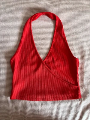 Röd ribbad omlott halterneck topp - Snygg röd ribbad halterneck topp med omlott framtill. Toppen är croppad och har djup v-ringning både fram och bak. Perfekt för dig som vill ha en trendig och bekväm topp till sommaren.