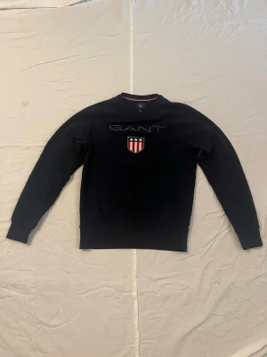 Svart sweatshirt från GANT - Stilren svart sweatshirt från GANT med rund halsringning och broderad logga samt ett rött och vitt sköldmärke med stjärnor på bröstet. Tröjan har ribbade muddar vid ärmslut och nederkant. Perfekt för en avslappnad och snygg look.