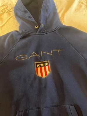 Blå hoodie från GANT med logga - Snygg blå hoodie från GANT med stor broderad logga och sköld med stjärnor och ränder på bröstet. Klassisk huva med dragsko och magficka framtill. Perfekt för en avslappnad och sportig stil.