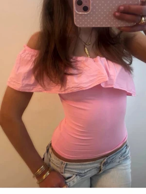 Rosa offshoulder topp med volang - Söt rosa offshoulder topp med bred volang runt axlarna. Toppen är ärmlös och har en croppad passform som ger en trendig look. Perfekt för varma dagar och somriga outfits.