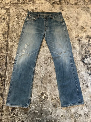 Blå raka jeans med slitningar - Säljer ett par blå raka jeans med slitningar och ljusa tvättade partier. Jeansen har klassisk femficksdesign, synliga slitningar på vänster lår och fransade benslut. Materialet är denim i bomull och passformen är relaxed.
