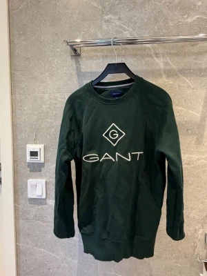 Mörkgrön sweatshirt från GANT - Mörkgrön sweatshirt från GANT med stor vit logga på bröstet. Tröjan har rund halsringning, långa ärmar och ribbade muddar vid ärmslut och nederkant. Perfekt för en avslappnad och stilren look. XS och S passar 