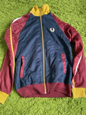 Retro ziptröja i blått och vinrött - Snygg retroinspirerad ziptröja från Fred Perry i marinblått och vinrött med gula detaljer. Tröjan har raglanärmar, ribbade muddar och krage i gult samt vita ränder på ärmarna. Broderad logga på bröstet och två fickor framtill.