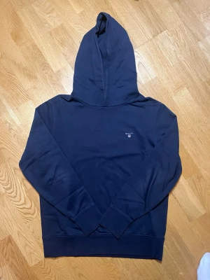 Mörkblå hoodie GANT - Säljer denna snygga mörkblåa hoodie från GANT i storlek M med liten broderad logga och sköld på bröstet. Klassisk modell med huva och ribbade muddar vid ärmar och nederkant. Tillverkad i mjuk bomullsblandning som känns skön mot huden. Perfekt för en avslappnad och stilren look. Vid frågor, extra bilder eller liknande är det bara att kontakta mig!