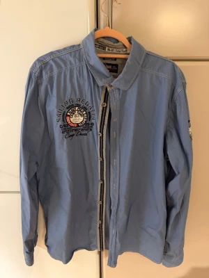 Blå skjorta från Camp David - Snygg blå skjorta från Camp David med broderad logga och text på bröstet. Skjortan har långa ärmar, klassisk krage och knappar framtill. Perfekt för dig som gillar detaljer och en sportig look.
