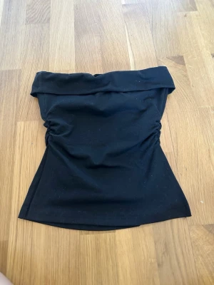 Svart tubtop med drapering - Snygg svart tubtop med lätt drapering i sidorna och en bred kant upptill. Toppen är ärmlös och har en figurnära passform, perfekt till jeans eller kjol. Mjukt och stretchigt material som känns bekvämt mot huden.