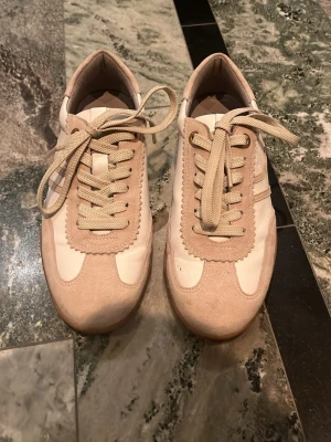 Beige och vita sneakers med mockadetaljer - Snygga sneakers i beige och vitt med vågiga mockadetaljer och klassisk snörning. Skorna har en retroinspirerad look och en låg silhuett. Perfekta för dig som gillar stilrena och neutrala färger. Skorna är köpta ifrån scorett och nypriset är 1400kr. Skorna är knappt använda och säljs pga fel passform.
