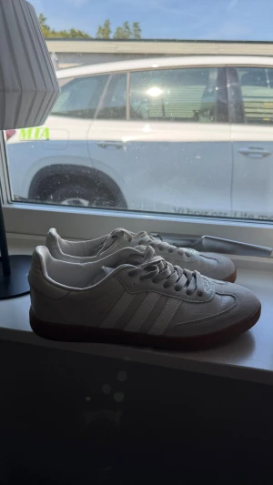 Vita Adidas Samba sneakers - Säljer ett par klassiska vita Adidas Samba sneakers med de ikoniska tre ränderna på sidan och brun gummisula. Skorna har lågt skaft och är tillverkade i läder med detaljer i mocka. Perfekta för en clean och tidlös stil. Använda ca 1 gång 