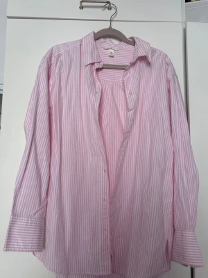 Rosa och vit randig skjorta - Säljer en klassisk långärmad skjorta med rosa och vita ränder. Skjortan har knappar framtill, krage och breda manschetter. Perfekt för dig som gillar en avslappnad men ändå stilren look.