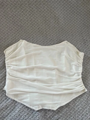 Vit ärmlös croppad topp - Säljer en stilren vit topp med croppad passform och ärmlös design. Toppen har lätt rynkade detaljer på sidorna och en mjuk, slät yta. Perfekt för dig som vill ha en enkel men snygg topp till sommaren.