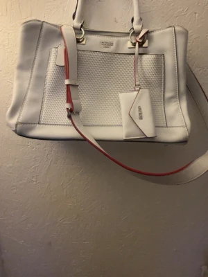 Vit handväska från Guess - Snygg vit handväska från Guess med silverdetaljer och strukturmönstrad framsida. Väskan har både handtag och en avtagbar axelrem med röda kanter samt en matchande liten accessoar. Perfekt för dig som vill ha en stilren och modern look.