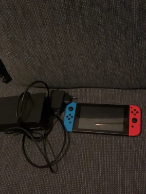 Nintendo Switch med blå och röd Joy-Con - Nintendo Switch spelkonsol med en blå och en röd Joy-Con-kontroll. Konsolen har en stor skärm och medföljer laddare och dockningsstation. Perfekt för gaming både hemma och på språng. Allt är i plast och elektronik med klassisk Switch-design.