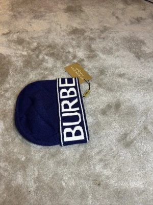 Blå mössa från Burberry - Snygg blå mössa från Burberry med bred vikt kant och stor vit logga framtill. Klassisk och stilren design som passar perfekt till kalla dagar. Mössan är stickad och har en mjuk känsla. Vet inte om real fick i present